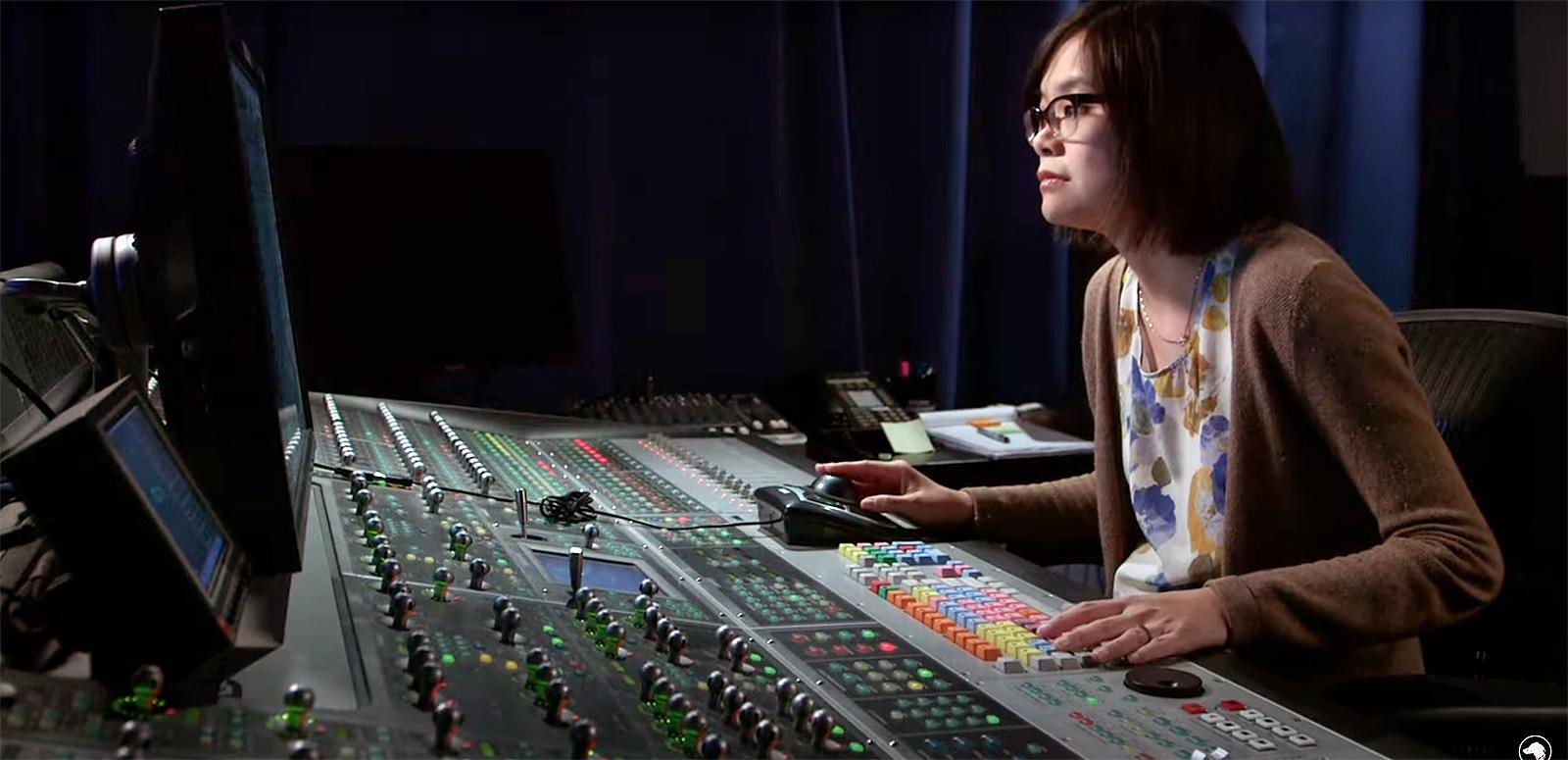 A woman sitting in a studio at a sound desk looking at a screen, there are a lot of buttons and dials in front of her. 