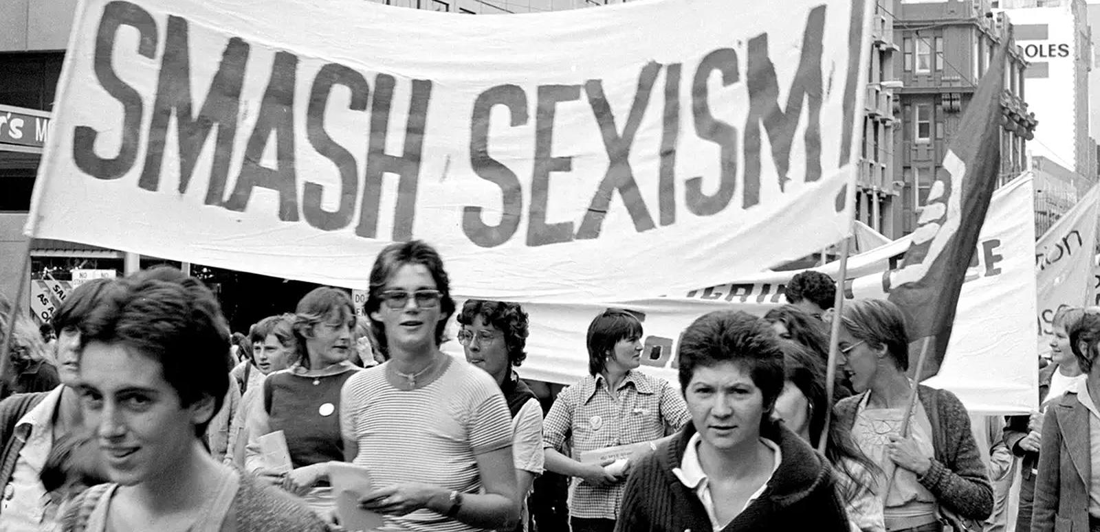 Women marching in the street with a big banner that reads SMASH SEXISM.