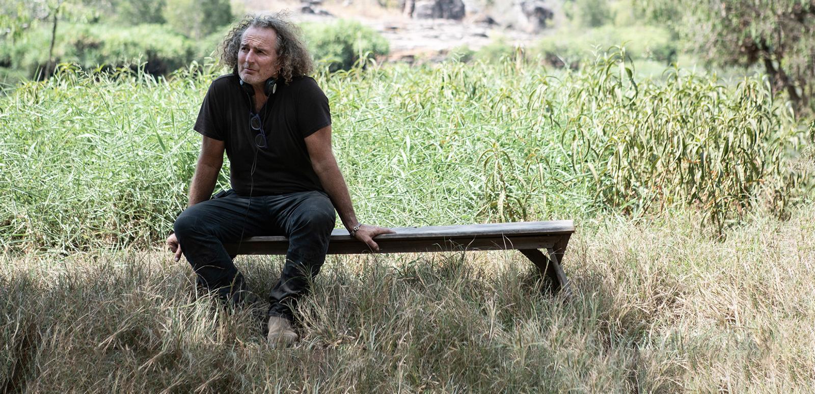 Director Stephen Maxwell Johnson sitting on a wooden bench in a grassy area on the set of his film High Ground. 