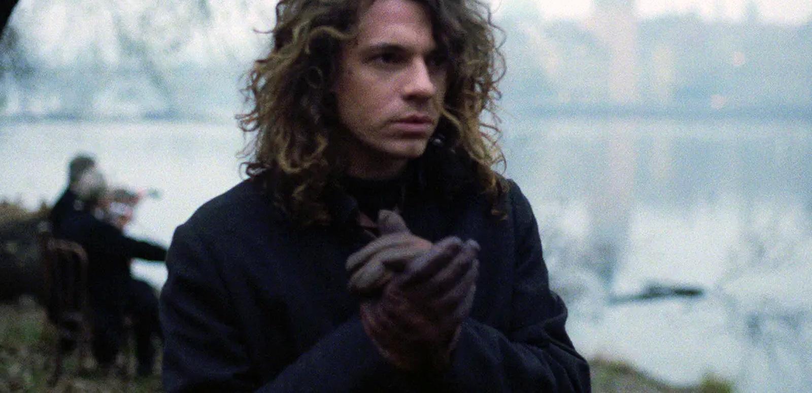 Head and shoulders shot of singer Michael Hutchence wearing a coat and gloves. There is a river and two people in the background.