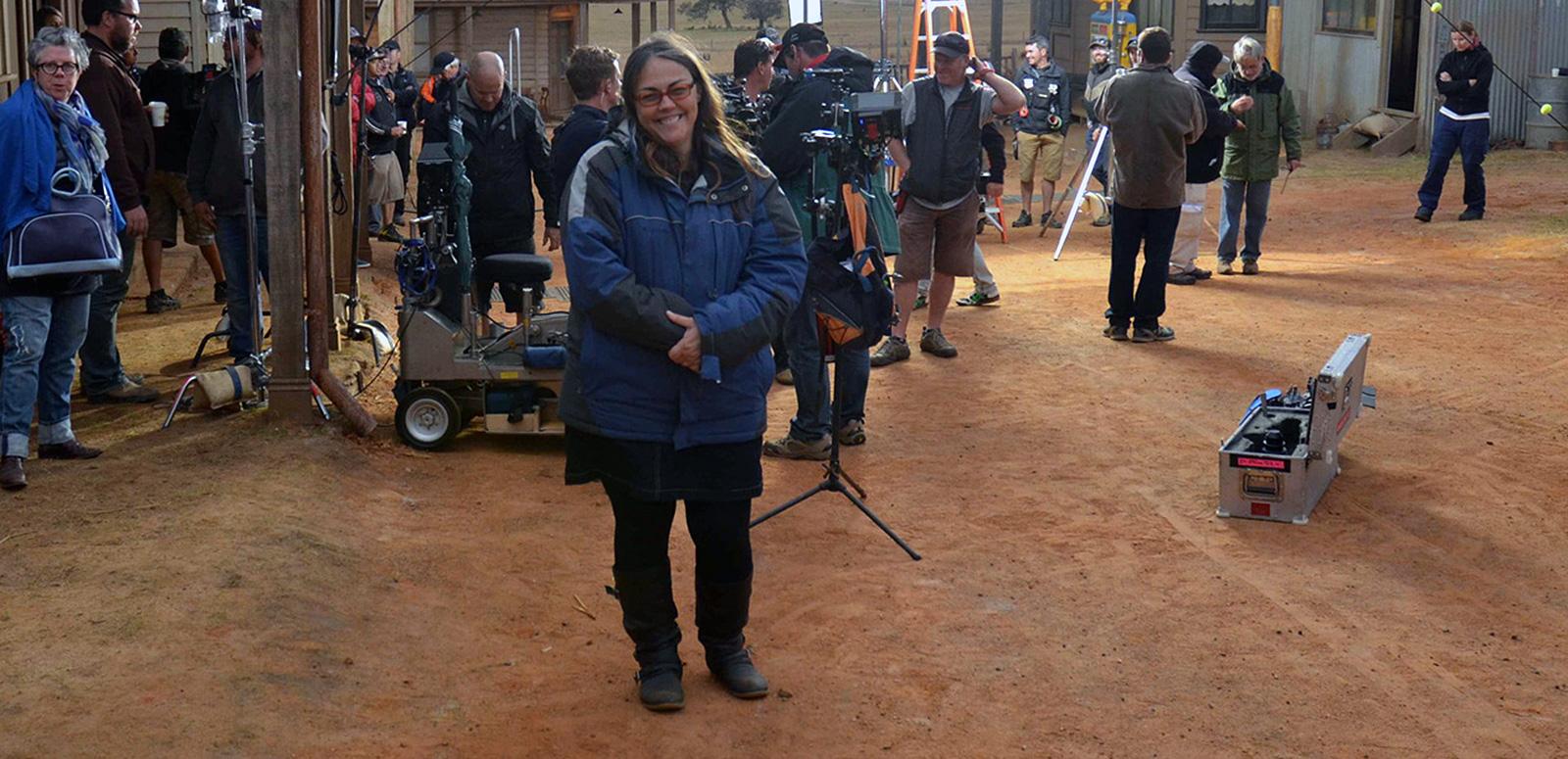 Director Jocelyn Moorhouse on the set of a film. She is looking at the camera while a large film crew are at work behind her. 