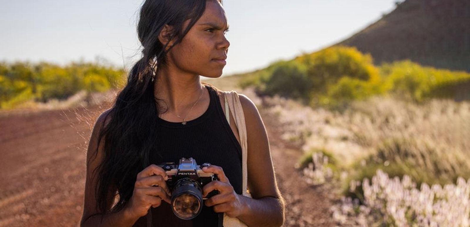 A teenage First Nations girl standing on an outback dirt road holding a camera and looking into the distance.