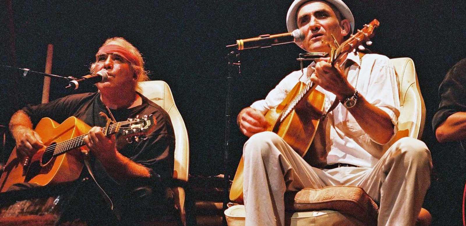 Kev Carmody and Paul Kelly on a stage seated in front of microphones and playing their guitars.