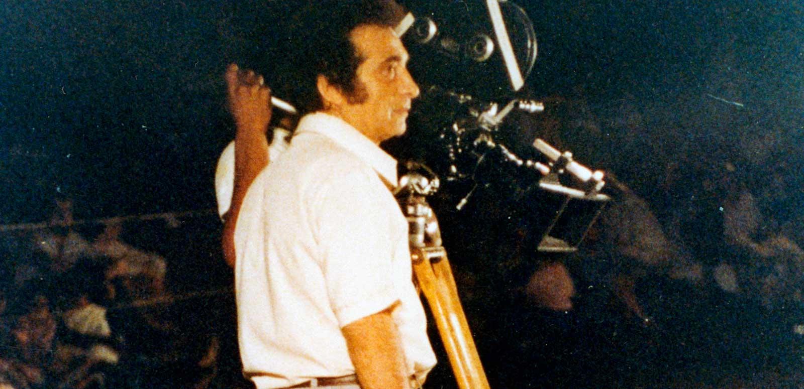 Filmmaker Giorgio Mangiamele standing outside in the dark next to a movie camera on a tripod. 