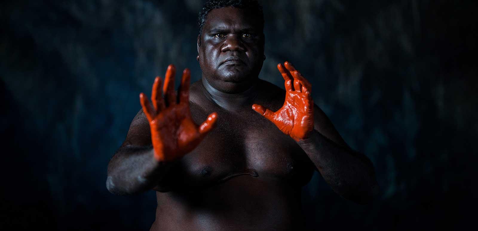 A First Nations man pictured from the waist up with his hands out in front of him. His hands are covered in red pain and he is looking directly at the camera. He is pictured against a dark background. 