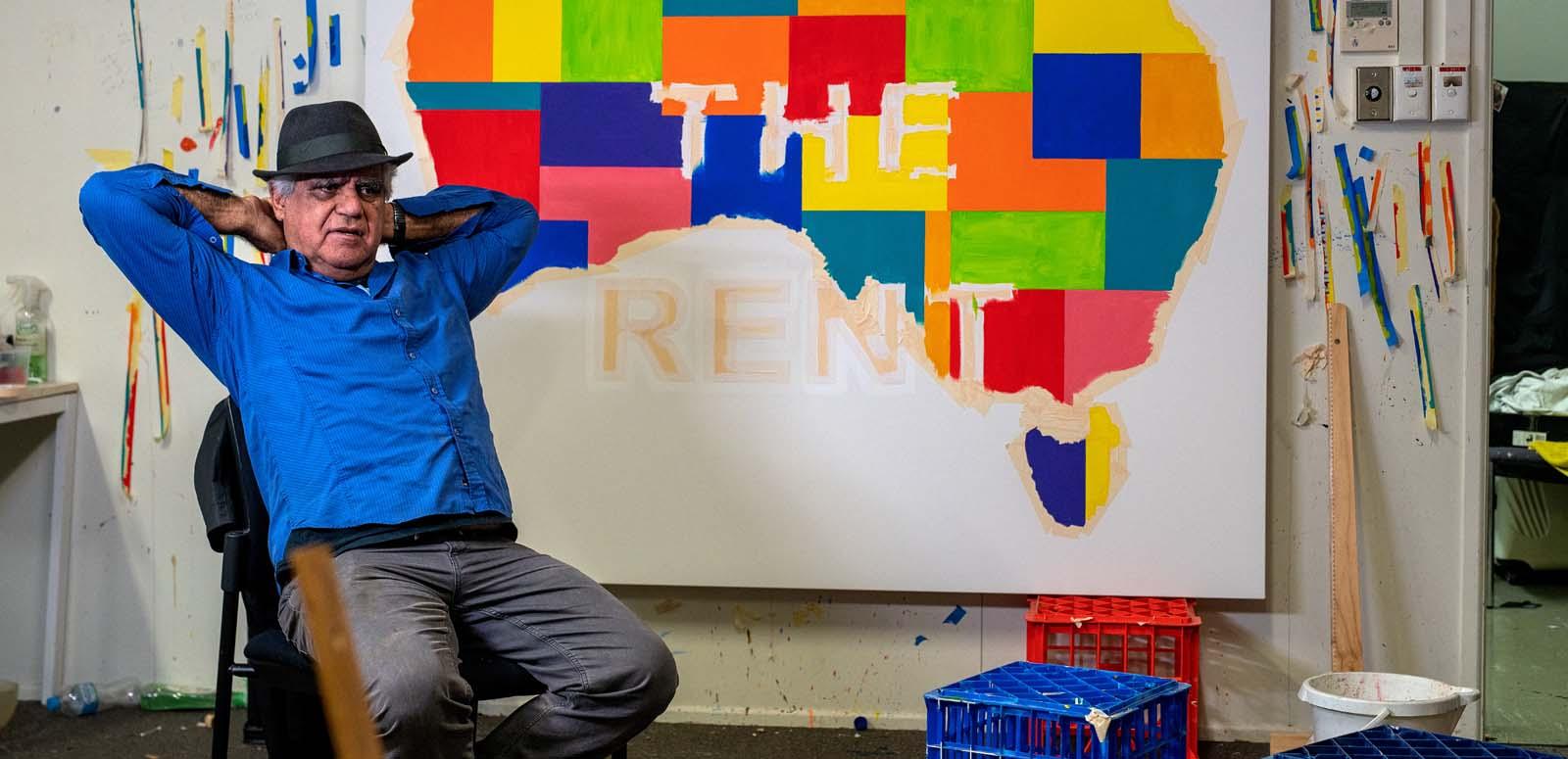 Artist Richard Bell seated in his studio in front of one of his works: a coloured Indigenous map of Australia with the words PAYE THE RENT across it