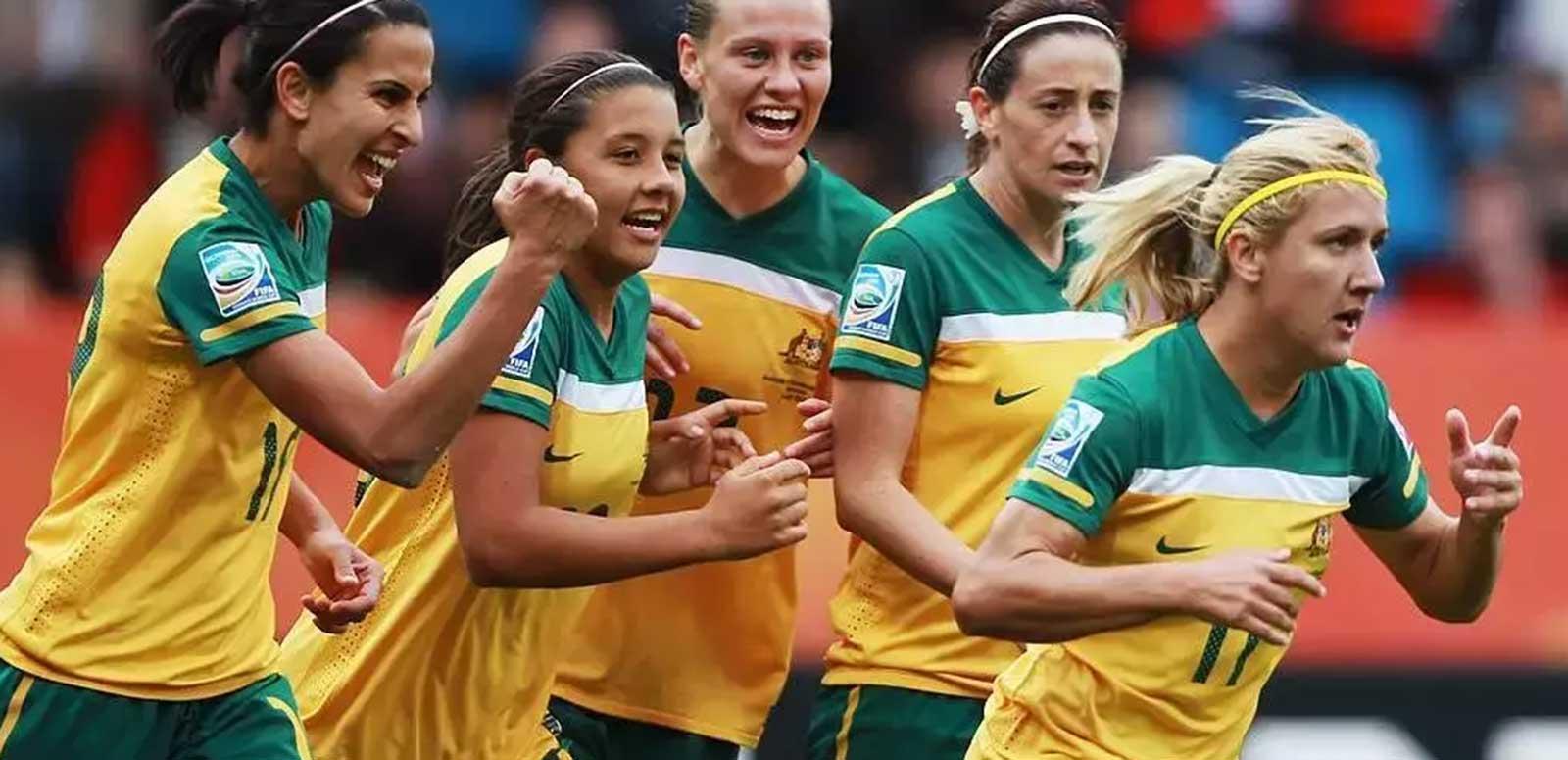 Five players from the Australian Women's Soccer team running side by side, they are smiling and look to be celebrating scoring a goal.