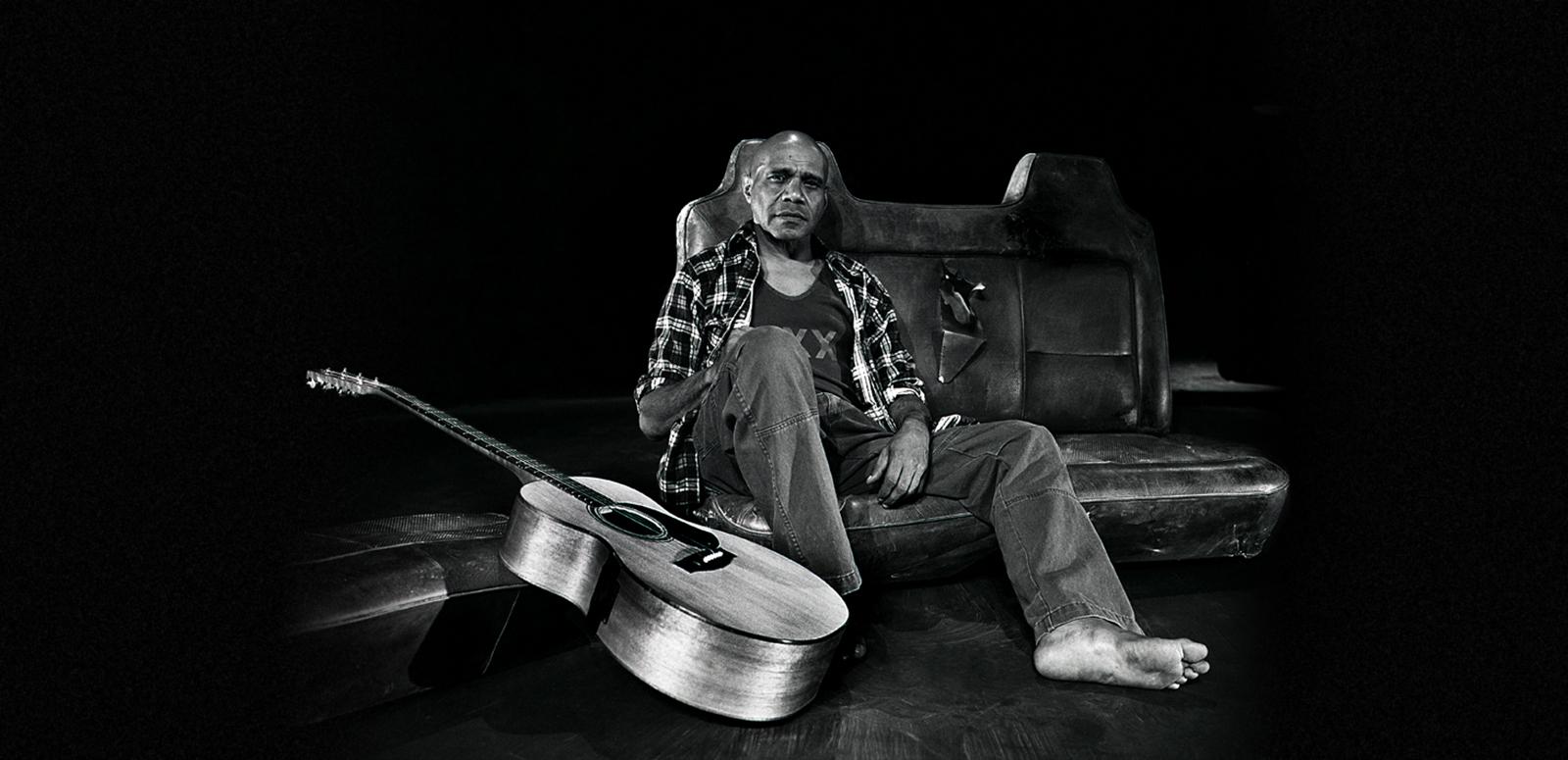 Archie Roach rests against an old car seat in a studio with his guitar resting nearby.