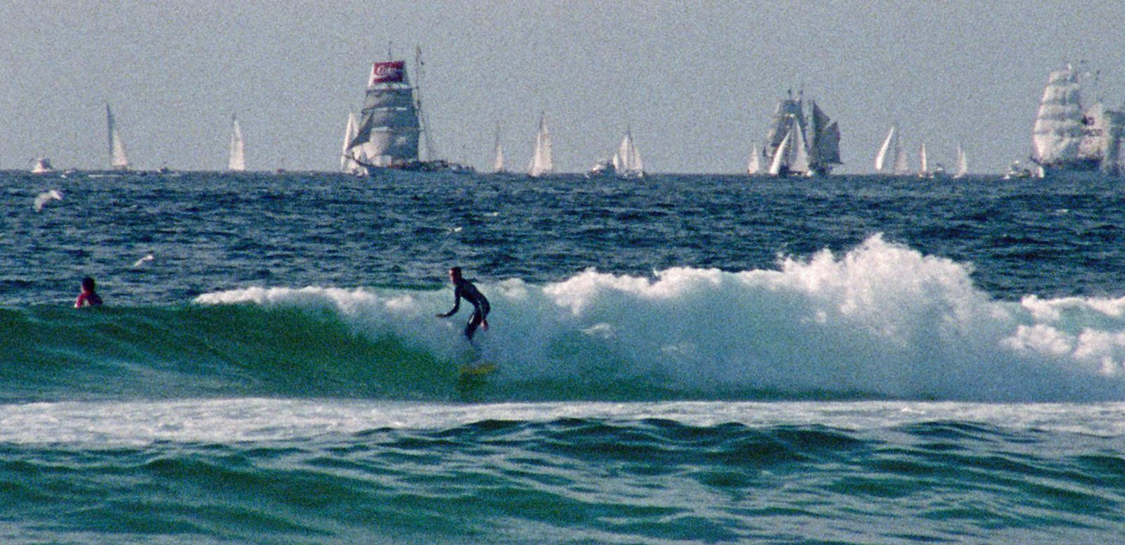 Two surfers in the ocean catching waves, in the background are tall ships and sailing boats.