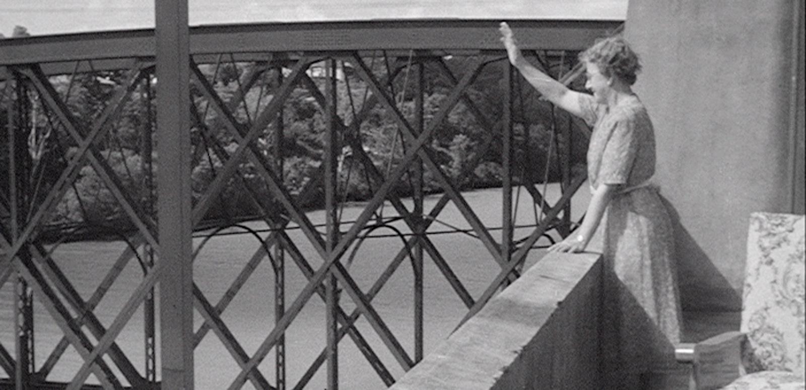 A woman is waving at someone we can't see. She is standing on a balcony with girders of a bridge nearby.