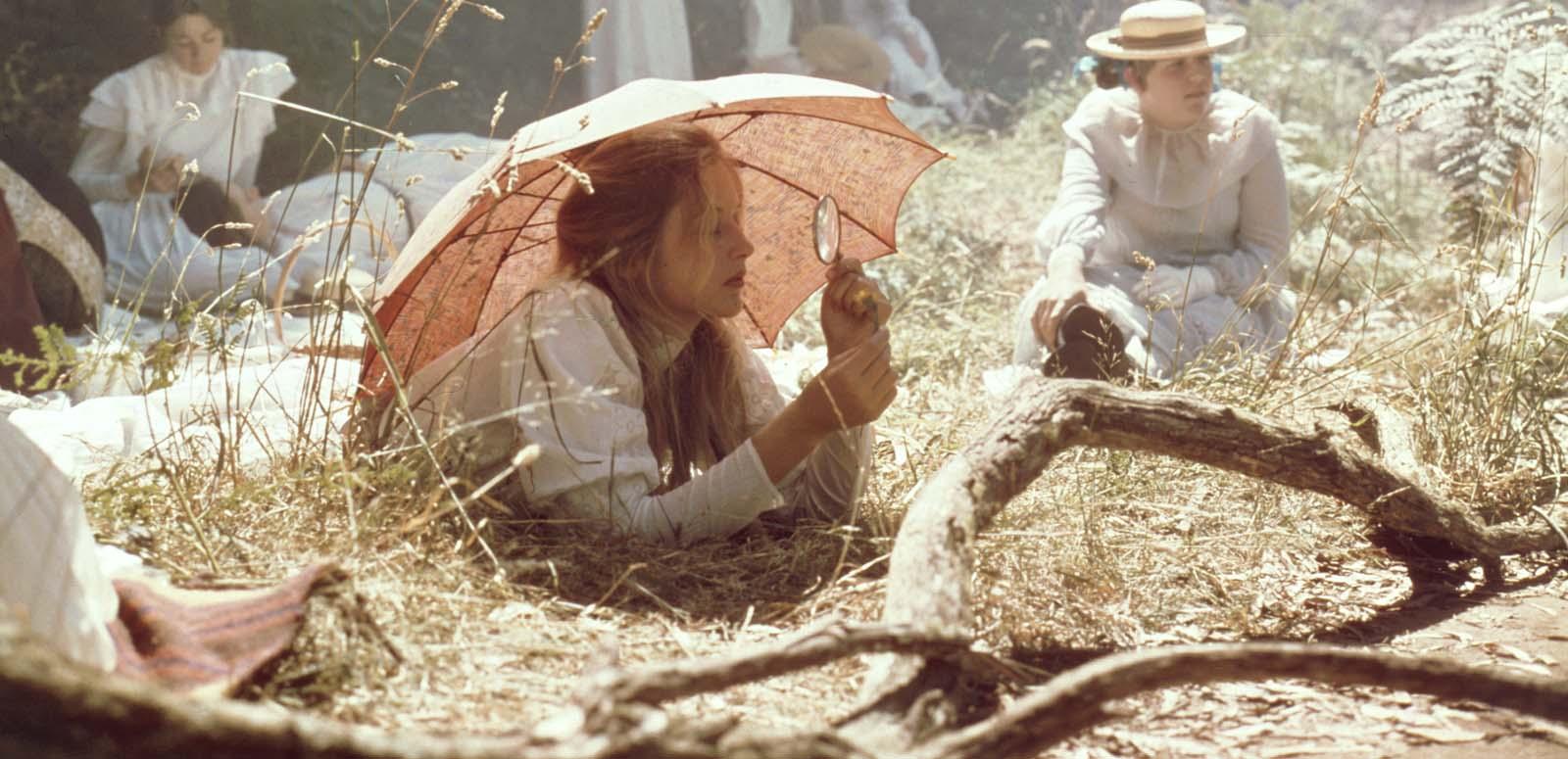 Miranda rests on the grass under a parasol and looks through a magnifying glass in a scene from the film Picnic at Hanging Rock