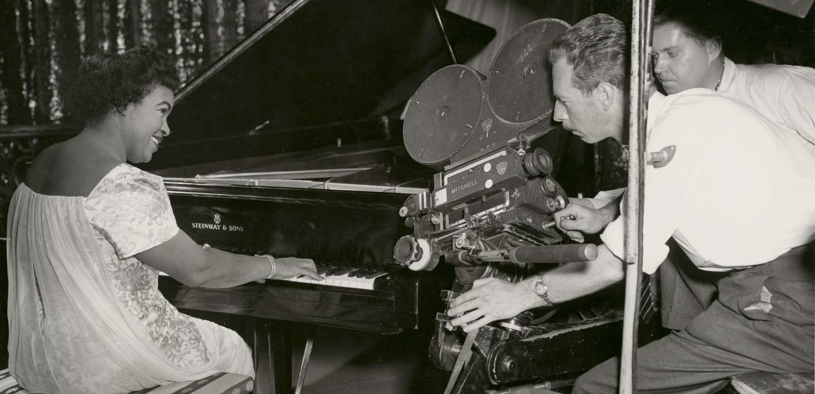 Winifred Atwell playing the piano and smiling while two men film her with a 35mm film camera next to the piano