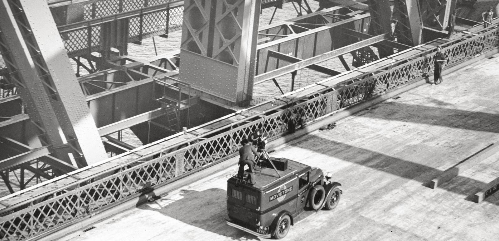 Fox Movietone Productions van with a man standing on top operating a camera. The van is on the deck of the newly-constructed Sydney Harbour Bridge.