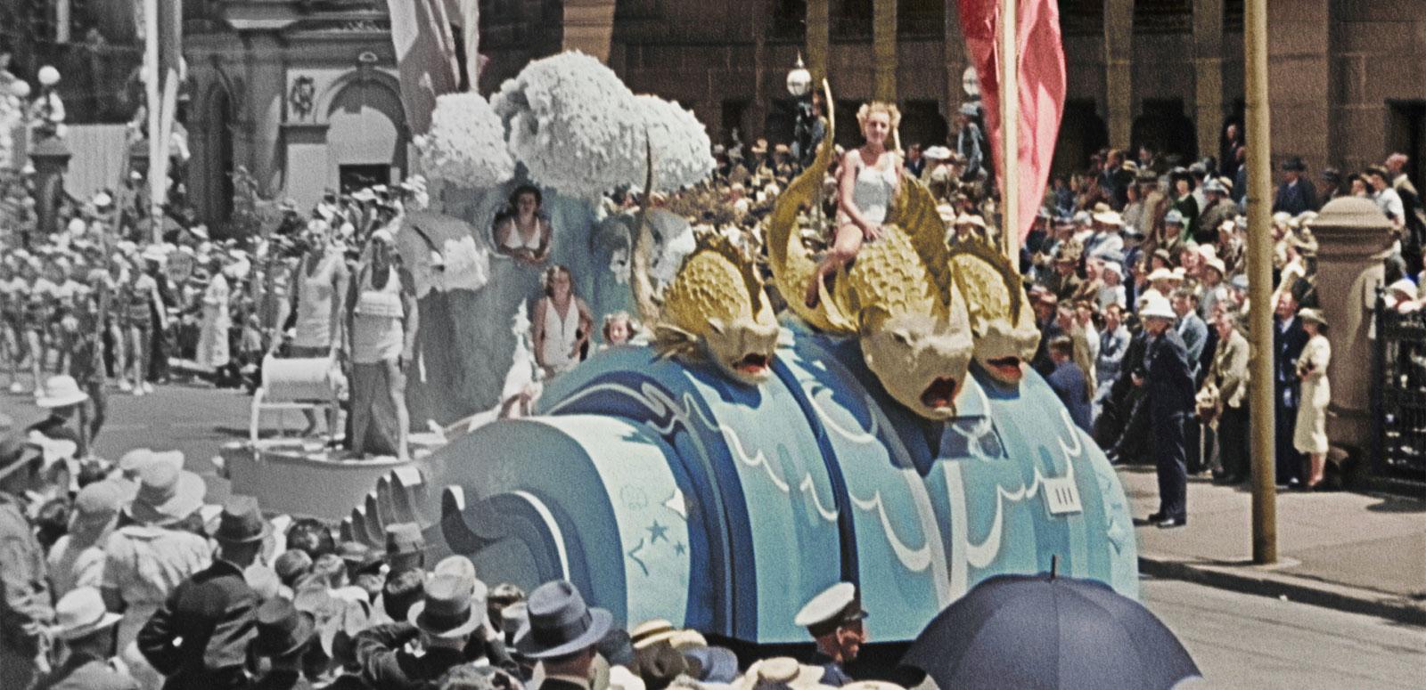 A partly colourised photo of a nautical-themed float in a 1930s street parade. On the float a woman dressed in white sits on a large-scale model of a sea creature.