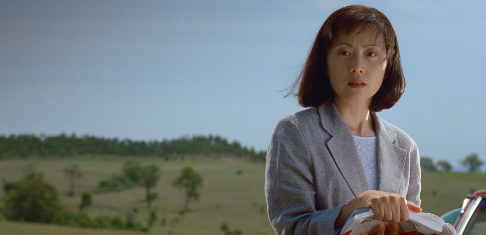 A young Asian woman dressed in a suit jacket pictured from the waist up in front of a green field and hills with trees in the background
