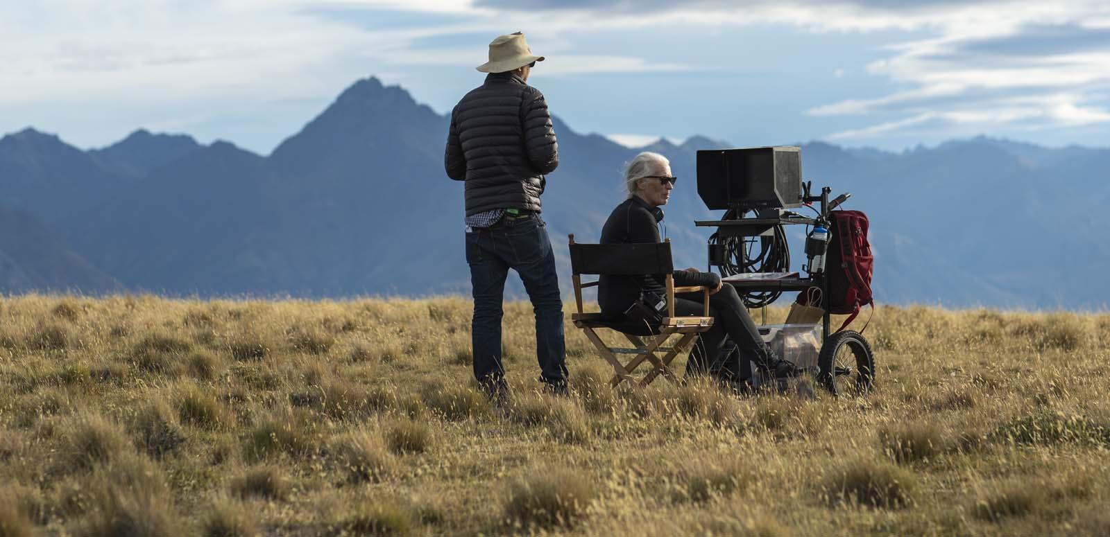 Cinematographer Ari Wegner standing next to director Jane Campion, seated at a monitor, on a grassy mountain location in New Zealand for the movie The Power of the Dog