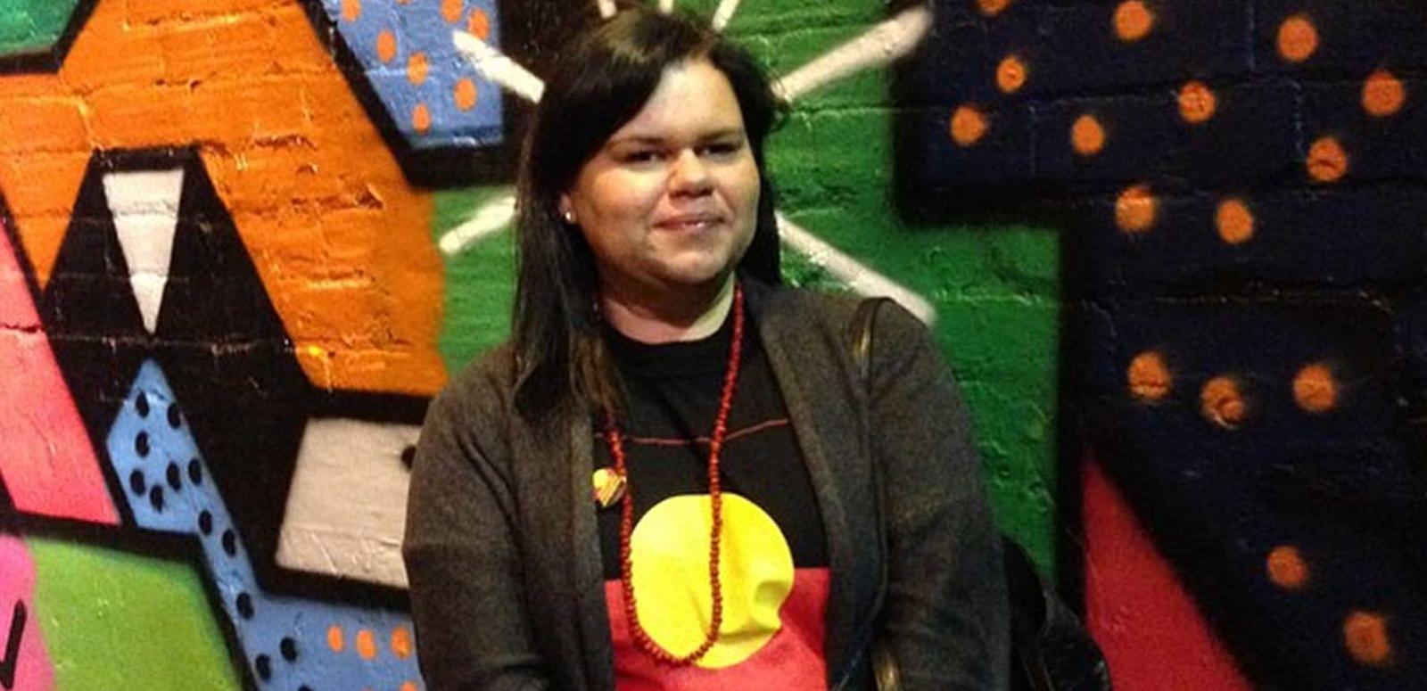 Tasha James standing in front of a colourful wall and wearing shirt bearing the Aboriginal flag