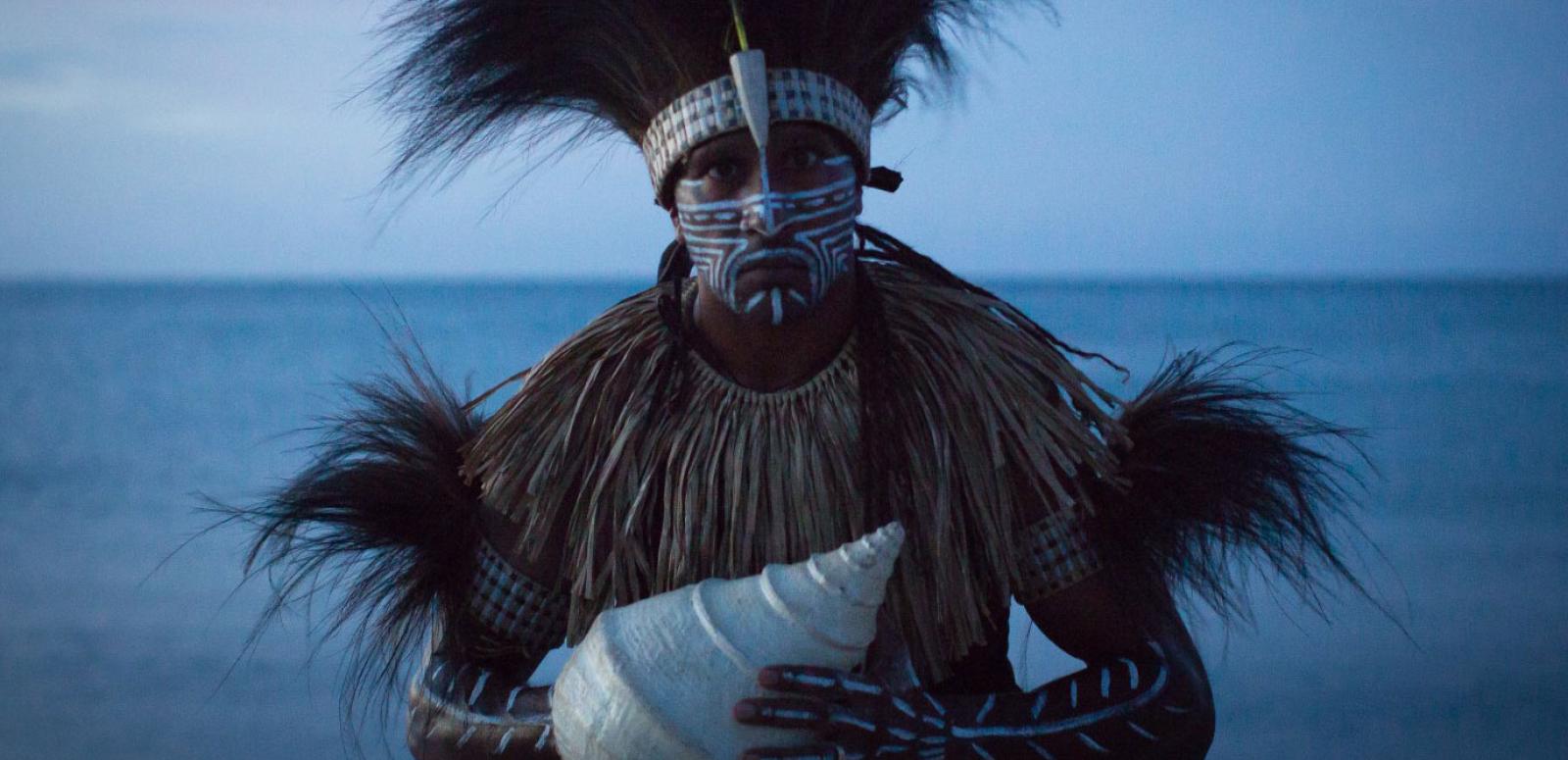 An indigenous man in traditional feathered costume holding a large shell with the ocean in the background.