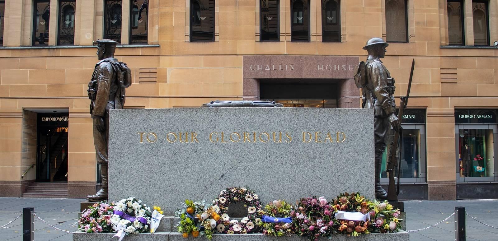 An Anzac Day memorial in Sydney inscribed 'To Our Glorious Dead' and flanked by statues of two soldiers. Wreaths of flowers surround the memorial