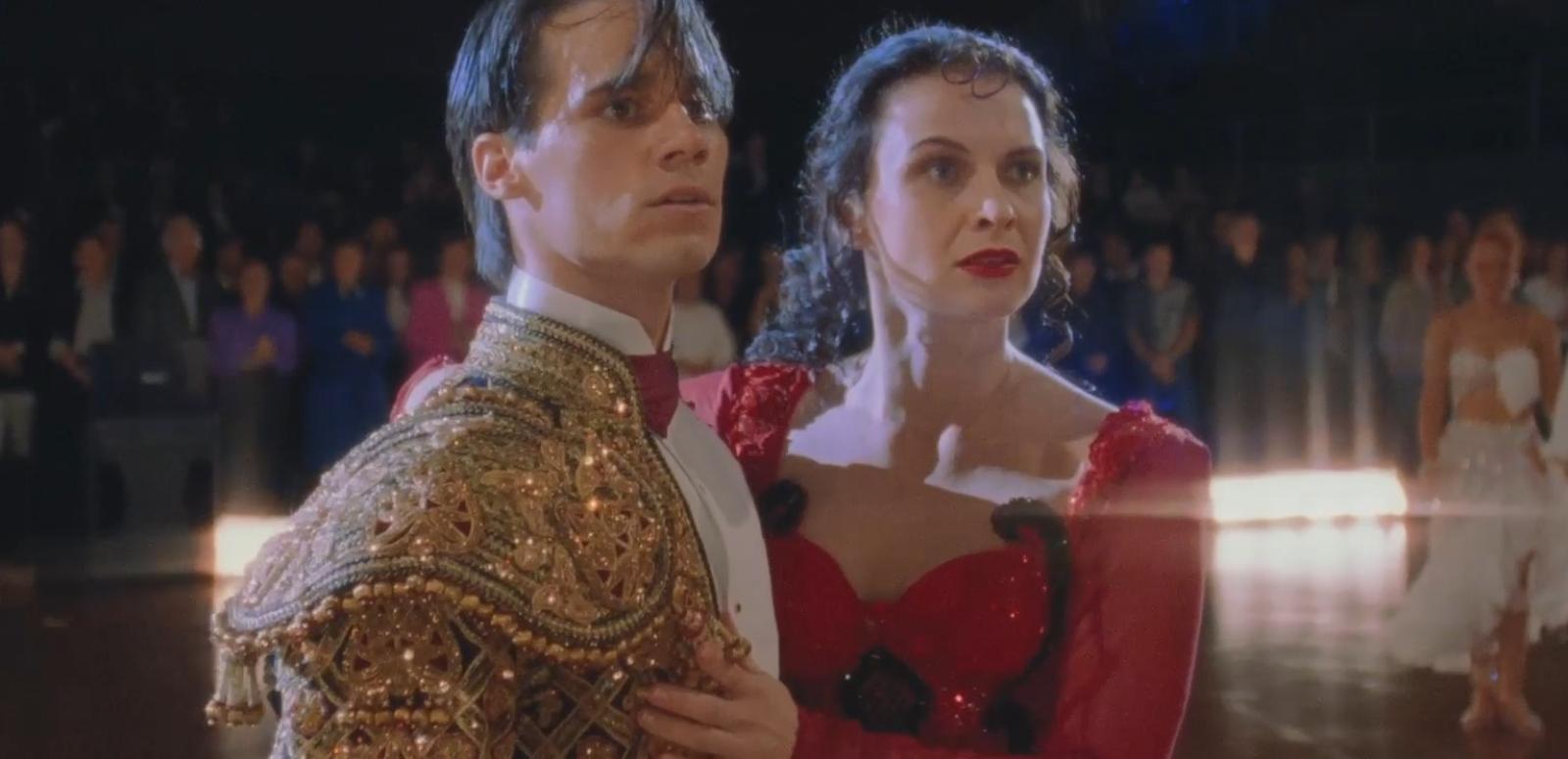 A young man and woman pictured from waist up on a dance floor wearing ballroom costumes look anxiously at something off camera