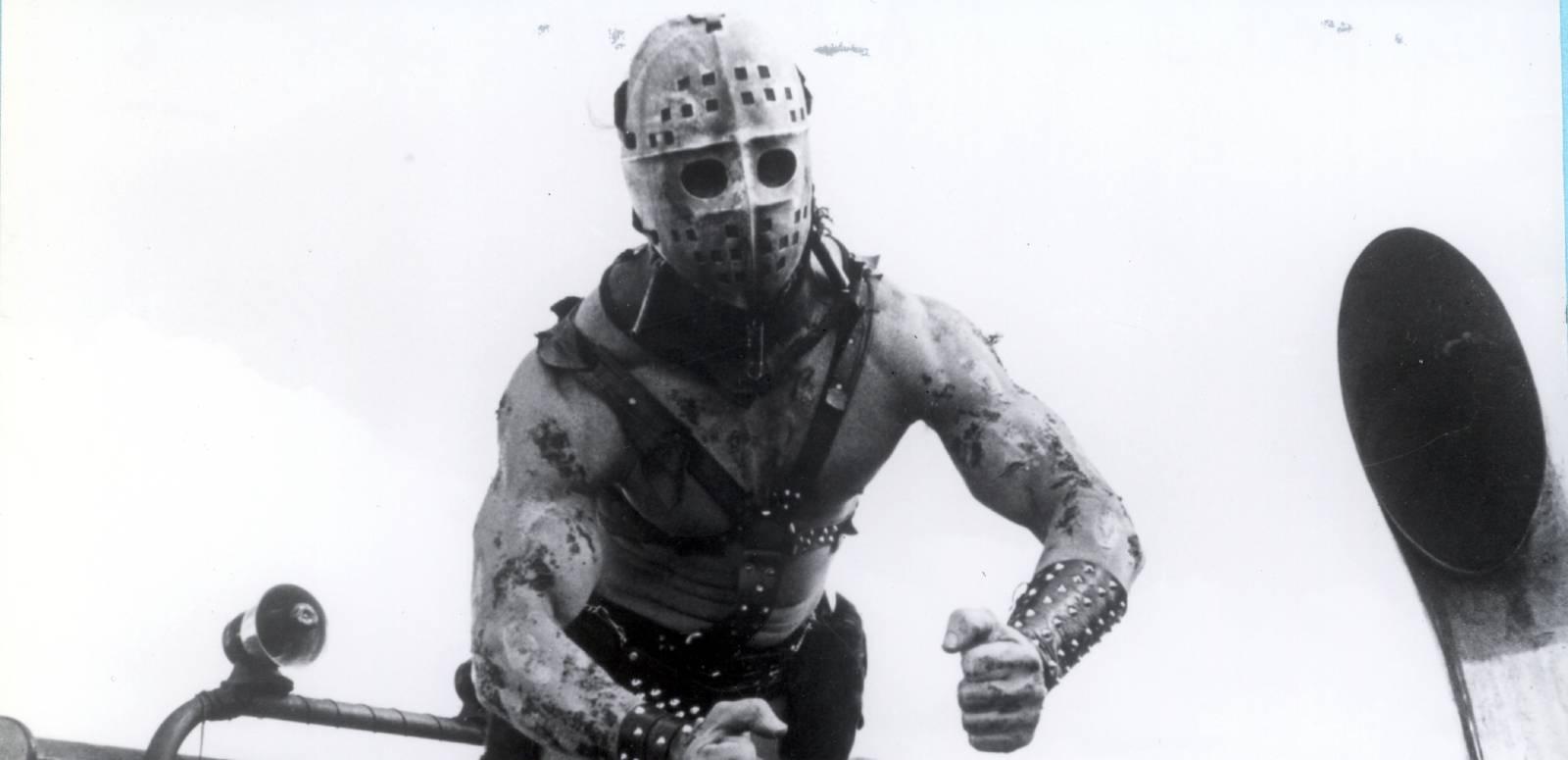 Mad Max 2 villain Humungus strikes a muscular pose with his face covered by a hockey mask