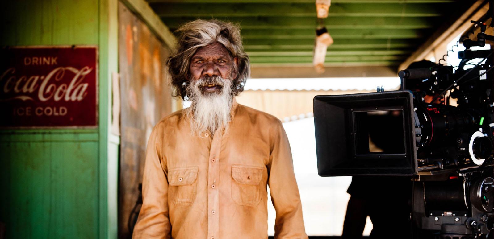 David Gulpilil on the set of Satellite Boy with a film camera beside him. He is looking directly at the camera.