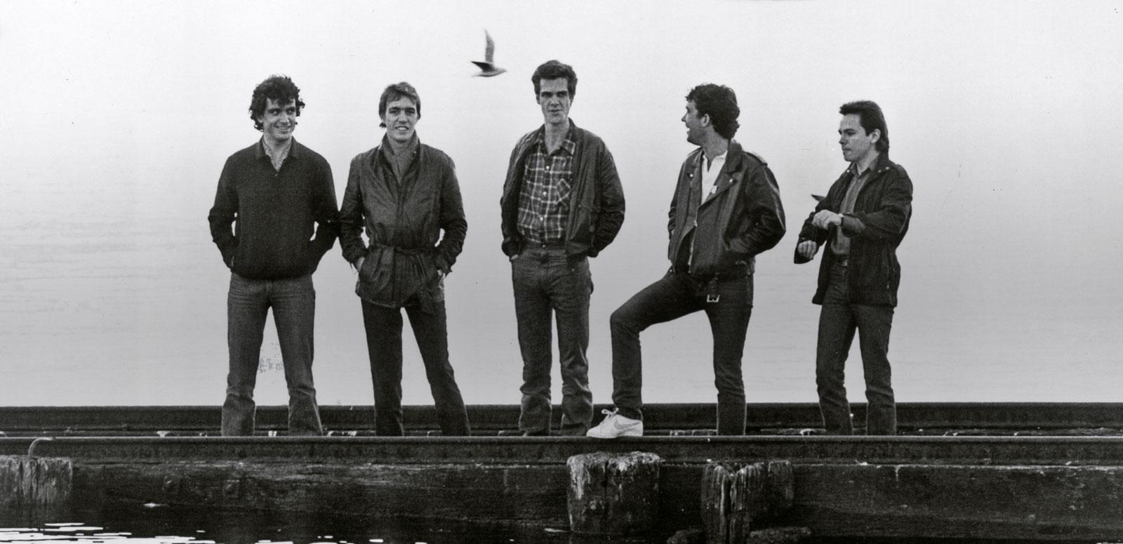 The five band members of Cold Chisel standing in a row on a pier with water in front and behind them. 