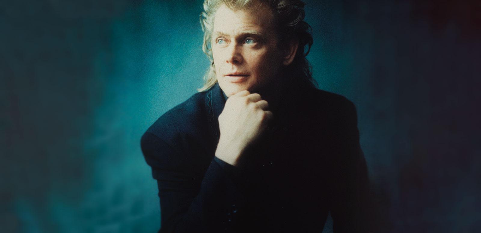 Image of John Farnham circa 1990 wearing a black jacket and seated with his chin resting in his hand.
