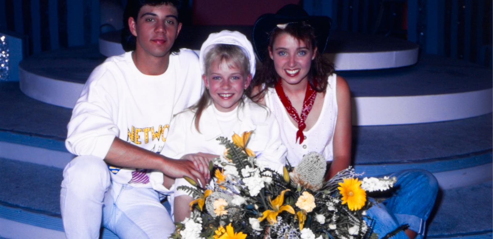 Young Talent Team members Vince Deltito, Rikki Arnot and Dannii Minogue sit together. Rikki, sitting in-between the two older teens, is holding a bunch of flowers.