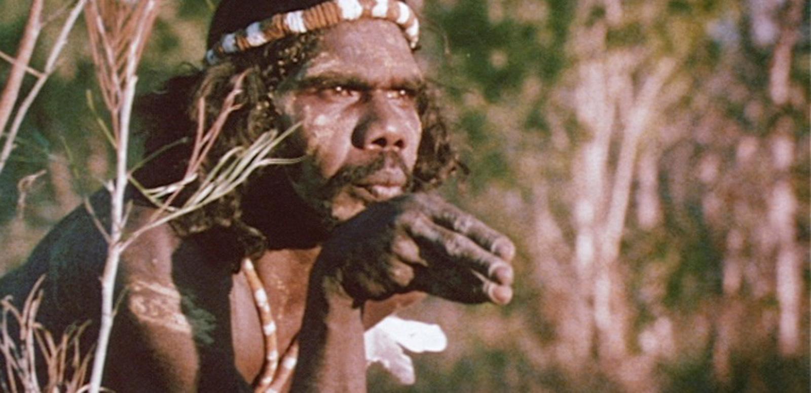 A young David Gulpilil is wearing traditional bodypaint in the bush. He is looking off-screen. He holds his hand in a dance pose in front of his mouth.