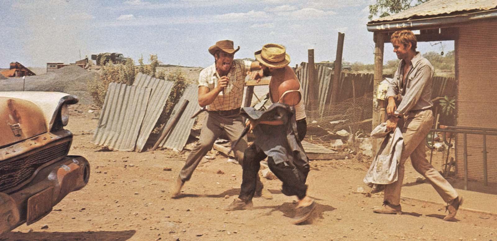 Jack Thompson and Donald Pleasence mock-brawling in the dusty outback while Gary Bond walks towards them