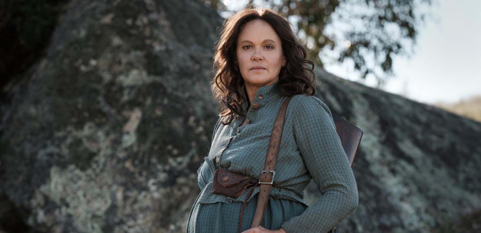 Leah Purcell as a pregrant bushwoman, standing outside in a rocky bush landscape in her film of The Drover's Wife