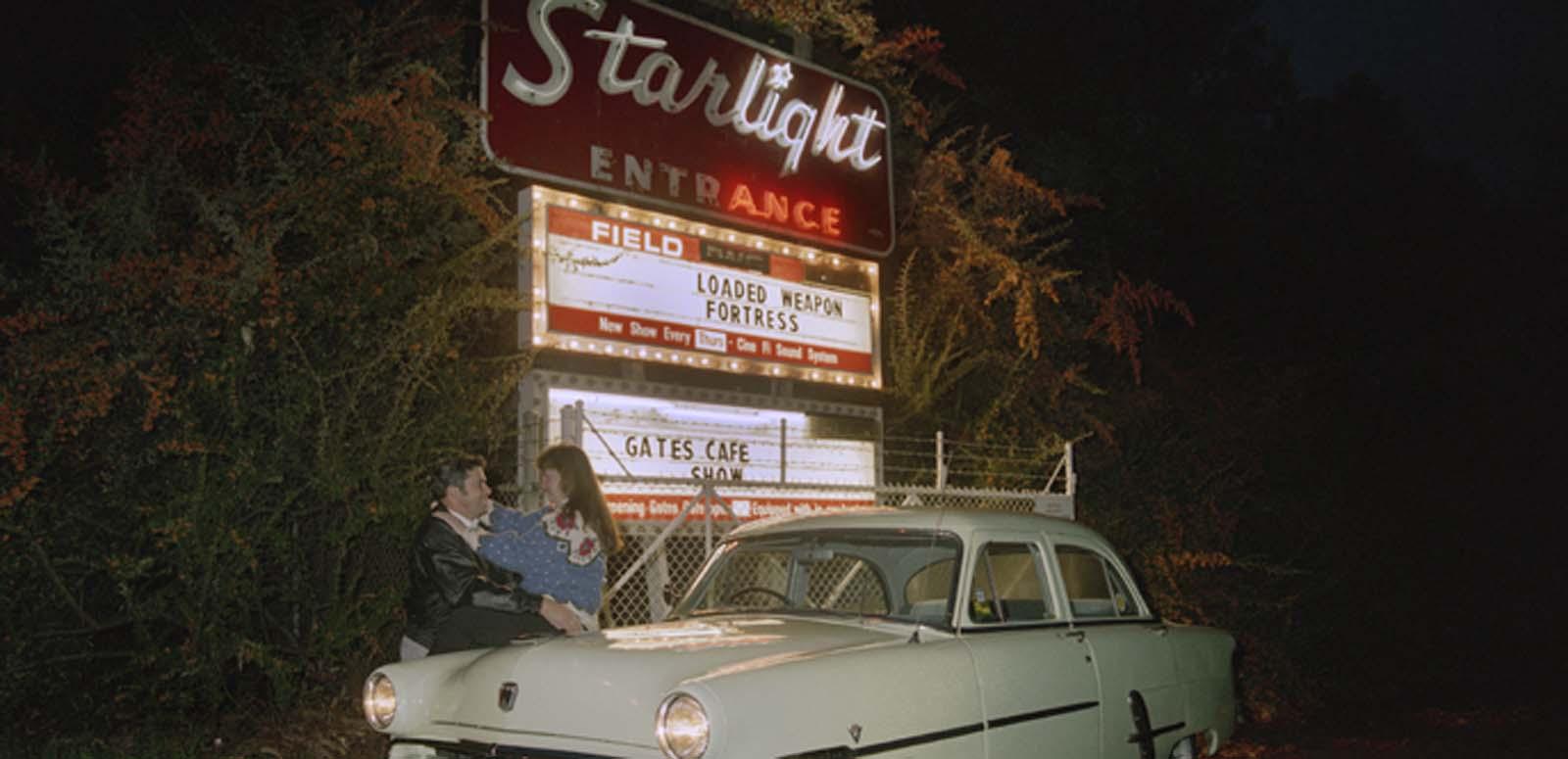 A man and woman embrace outside a drive-in cinema at night, leaning against their car