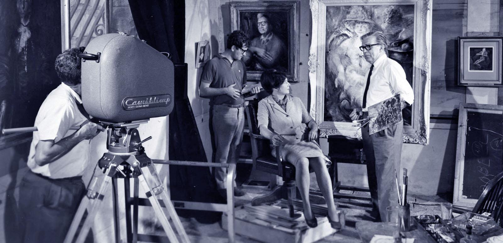 Blue-tinted black and white still of a man is operating a large film camera. He is filming artists Margaret Olley and William Dobell. There are two portraits painted by Dobell in the background as well as another member of the film crew.