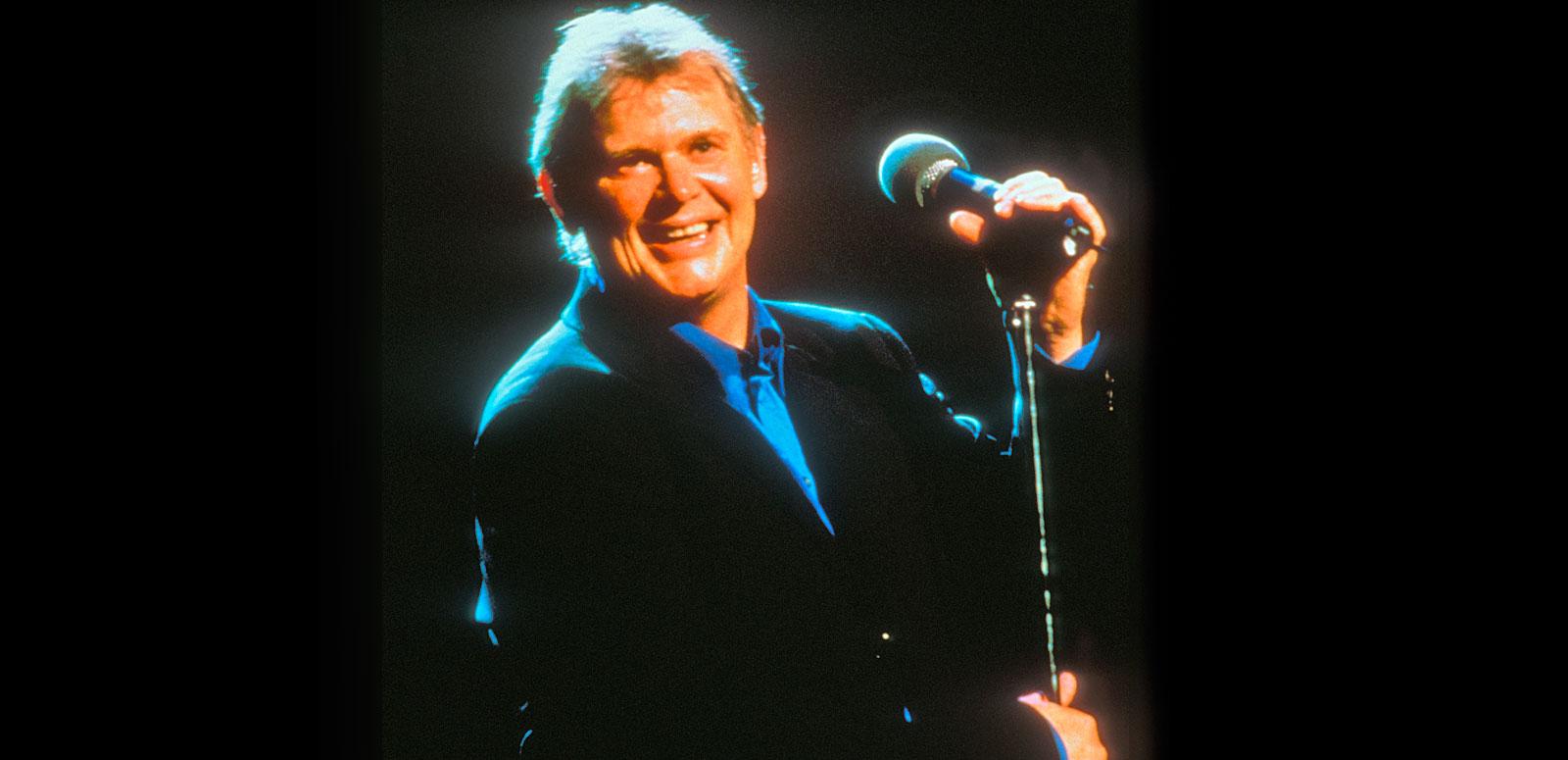 Singer John Farnham, circa late 1990s or early 2000s, pictured from the waist up wearing a black jacket and blue collared shirt, standing on a stage and holding on to a microphone stand. He's smiling at the camera.