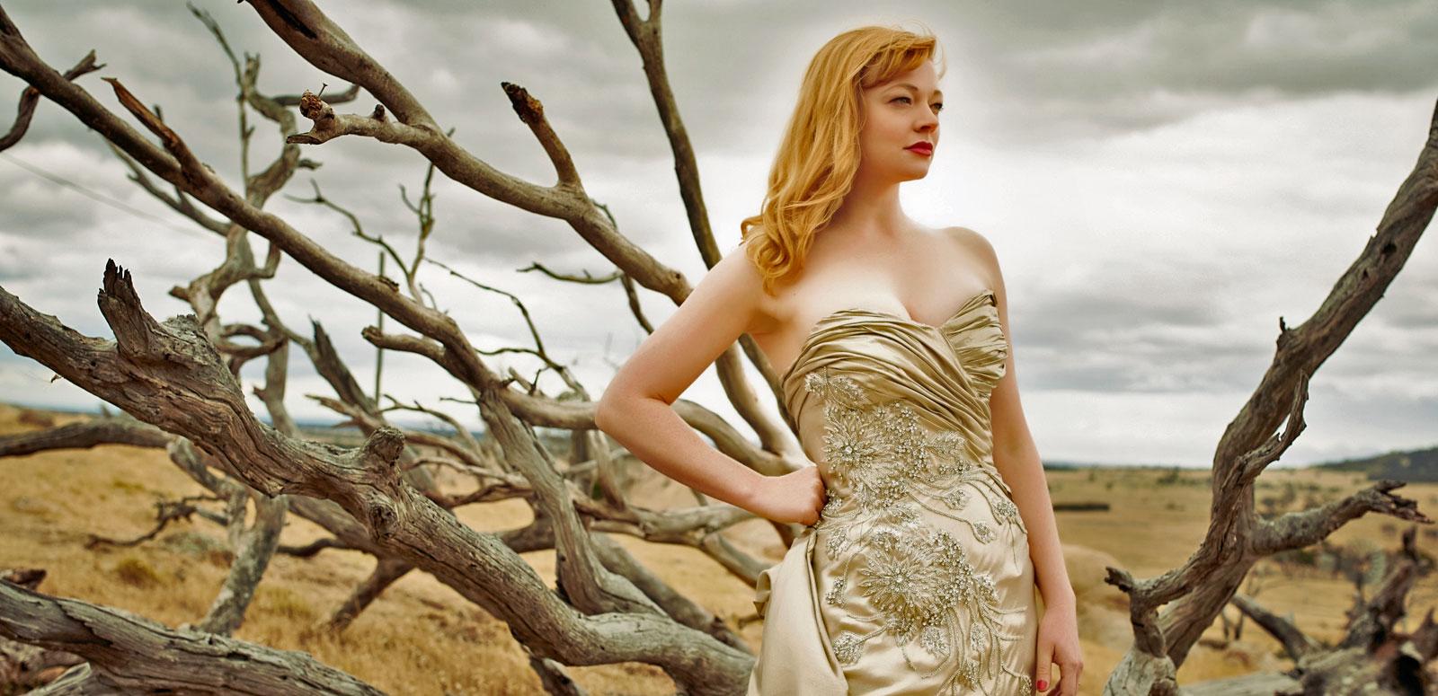 Actress Sarah Snook in a publicity shot for The Dressmaker, wearing a fitted, champagne-coloured, beaded gown and standing in front of some bare tree branches. In the distance you can see a brown, dusty plain and grey, cloudy sky.