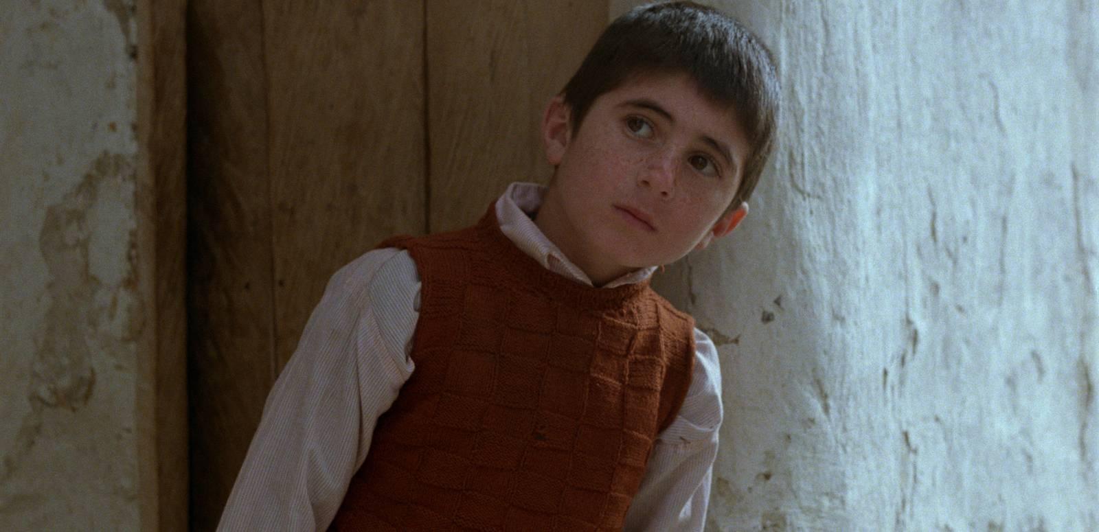 A young boy in collared shirt and vest stands in front of a closed wooden door, looking quizzical
