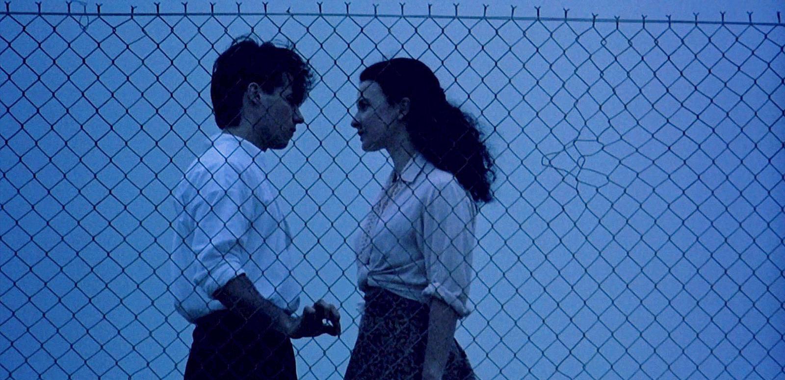 Paul Mercurio and Tara Morice in scene from the film Strictly Ballroom. They are facing each other in front of a wire fence. The time of day seems to be early evening. 