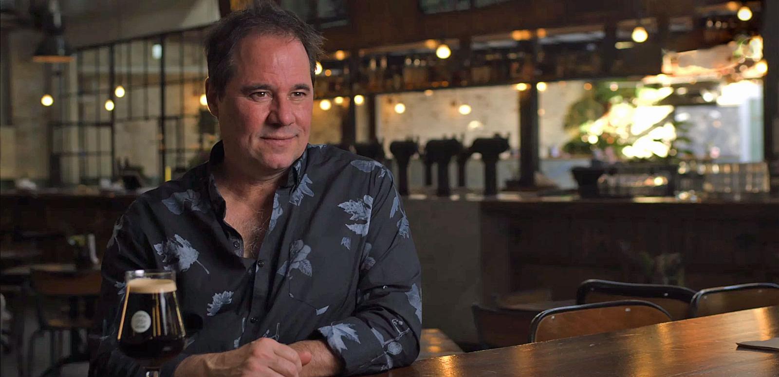 Waist-up shot of actor Paul Mercurio sitting at a restaurant table with a bar in the background. 