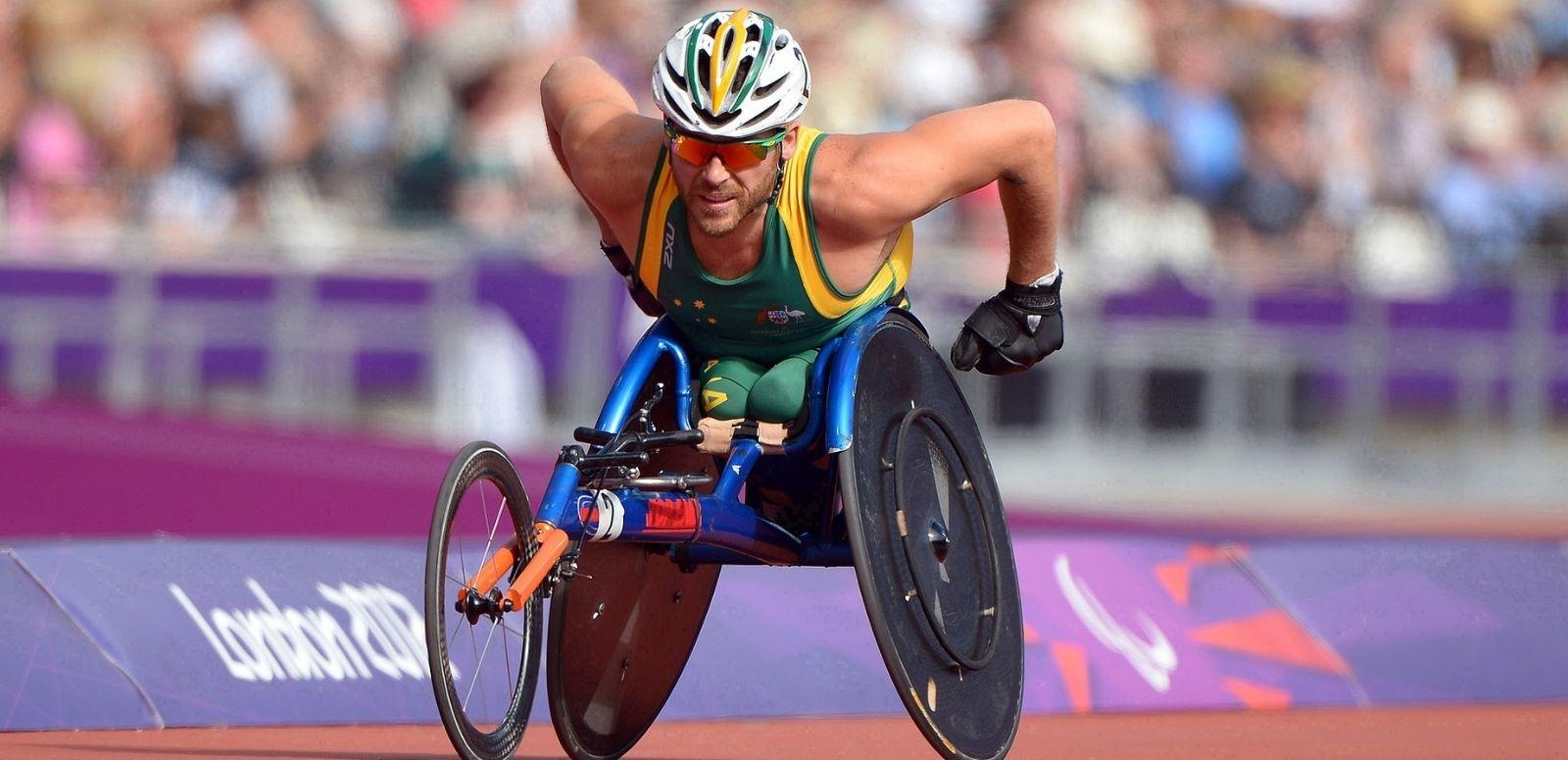 Wheelchair racer Kurt Fearnley is captured racing at the London 2012 Paralympics. A packed stadium is blurred behind him.