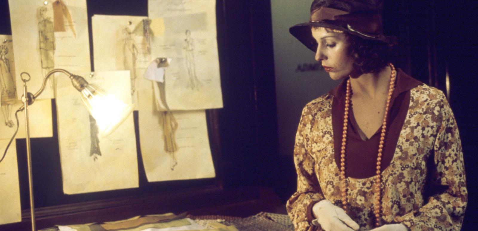 Film still of Helen Morse as Caddie looking at drawings of women's dress designs in a studio.