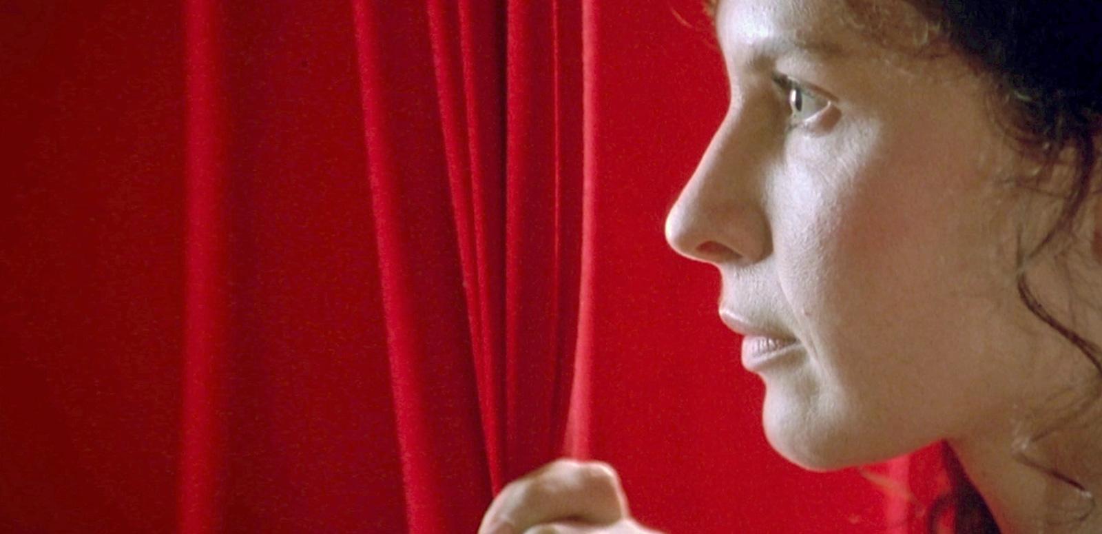 Close up of Tara Morice in profile as her character Fran in the film Strictly Ballroom. Fran his pulling aside a red curtain and looking past it, facing left of camera.
