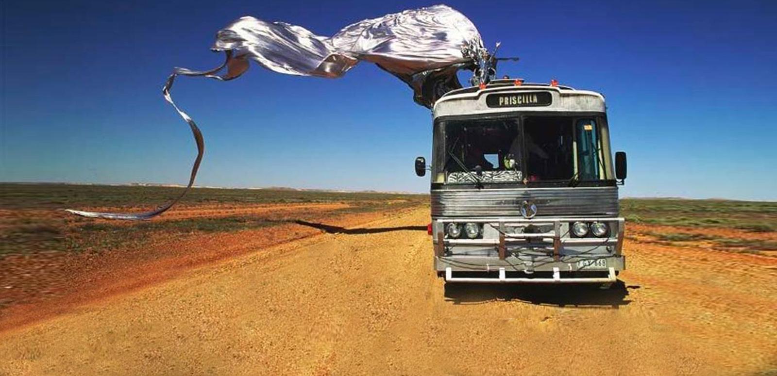 Scene from Priscilla, Queen of the Desert. A tour bus on a dirt road in the desert with blue sky in the background. On top of the bus is Guy Pearce in a shiny silver frock sitting on a giant stiletto shoe with a long train flowing behind the bus.