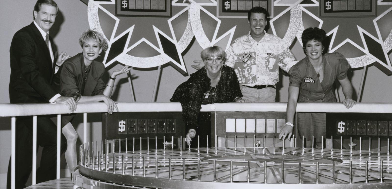 Game show hosts and contestants in a TV studio standing behind a big spinning wheel.