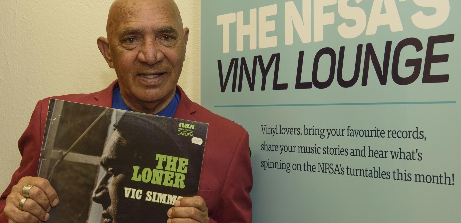 Vic Simms holding a copy of his record The Loner in front of the Vinyl Lounge banner
