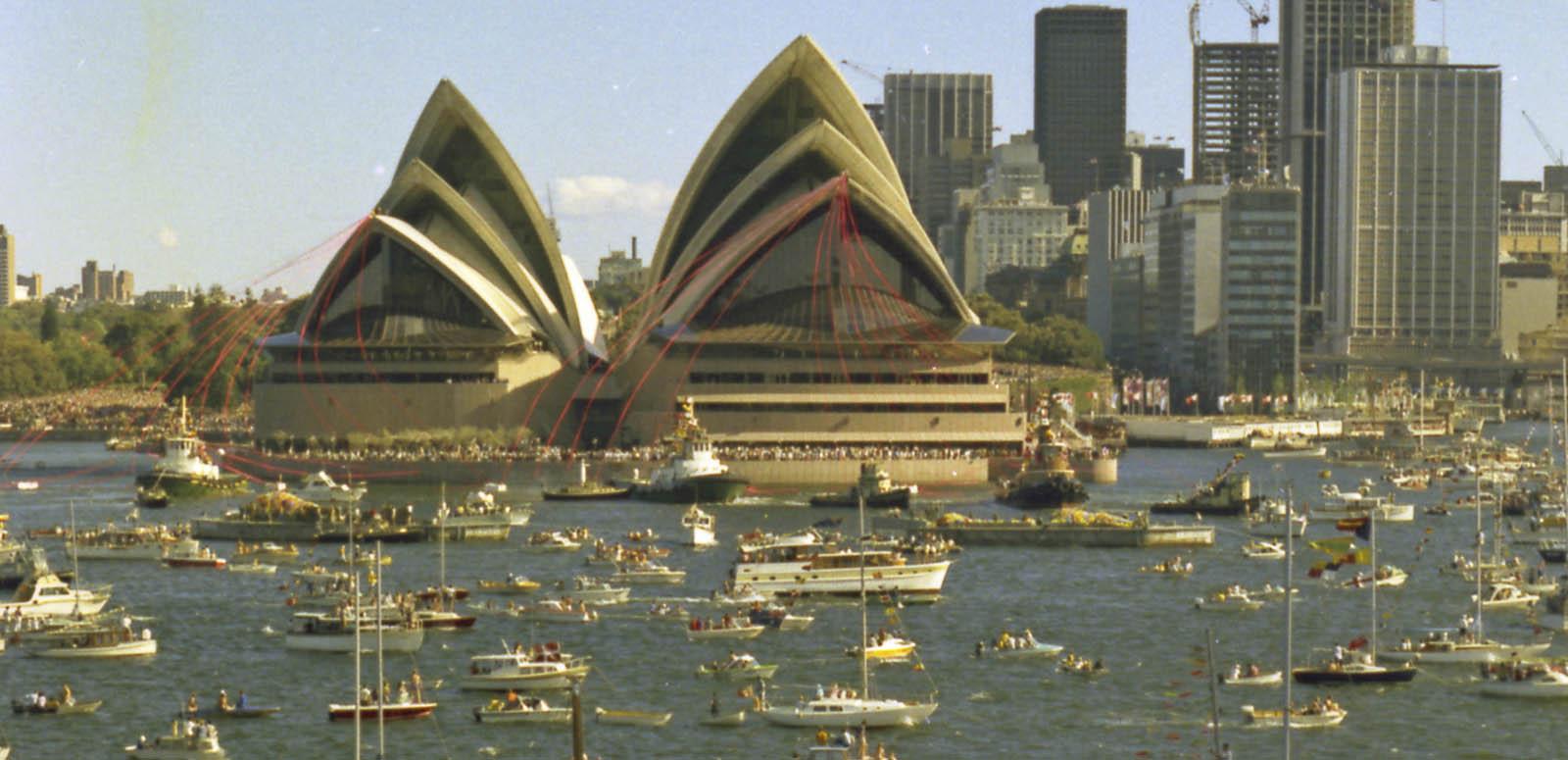 The Sydney Opera House is festooned with red ribbons. There are many boats on the water in front of it.