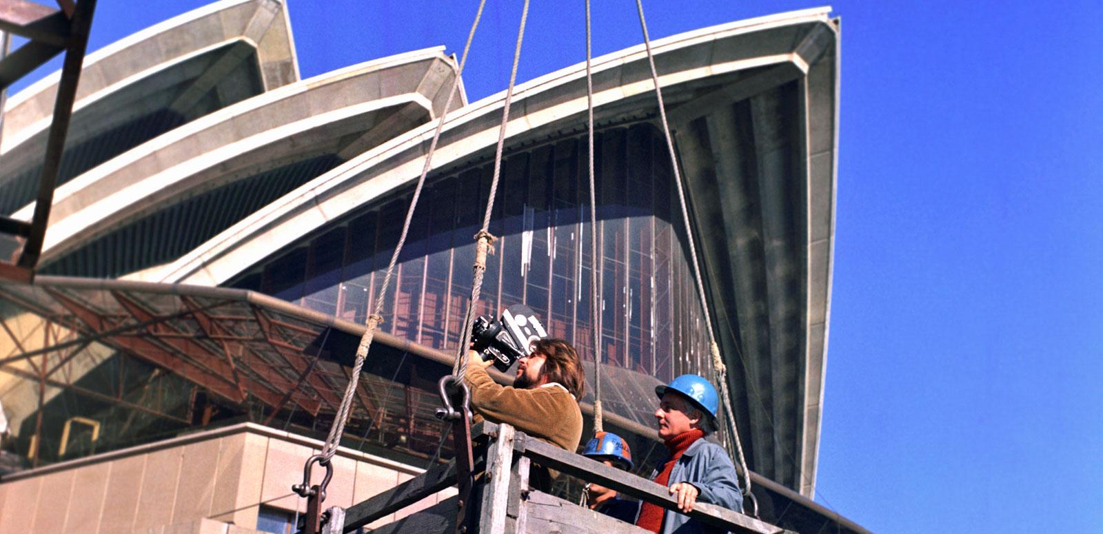 The sails of the Opera House tower above a camera operator and a man in a hardhat. They are standing in a wooden box which is hoisted to a crane.