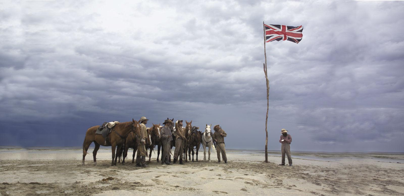 A group of men and horses standing on a plain in the Australian outback stand looking at a Union Jack flag that has been raised on a long, thin tree trunk.