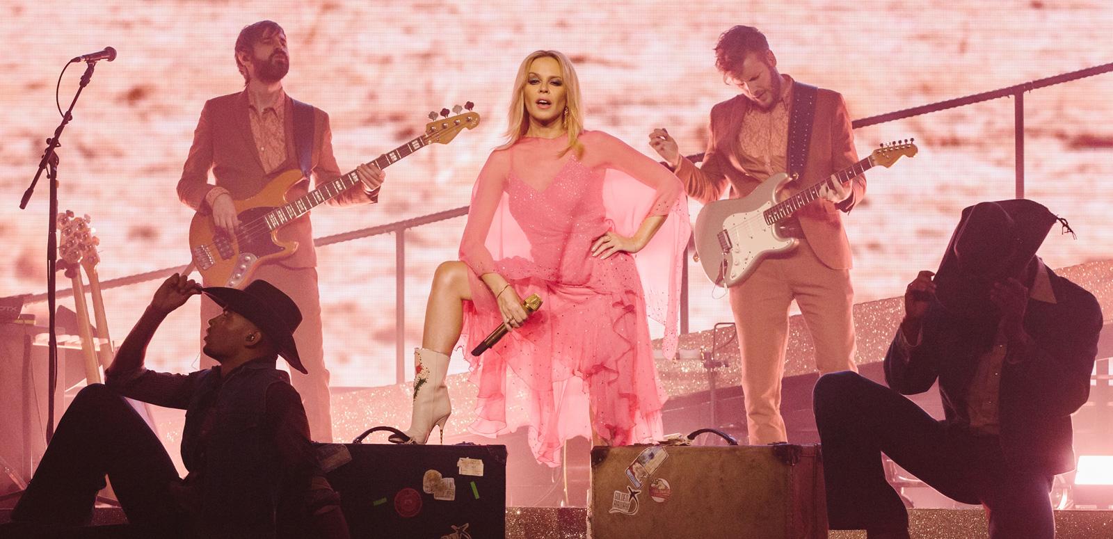 Kylie Minogue is standing on a stage with a foot up on an amplifier in a power pose. There are dancers in front of her and two musicians on stage with her playing guitar.