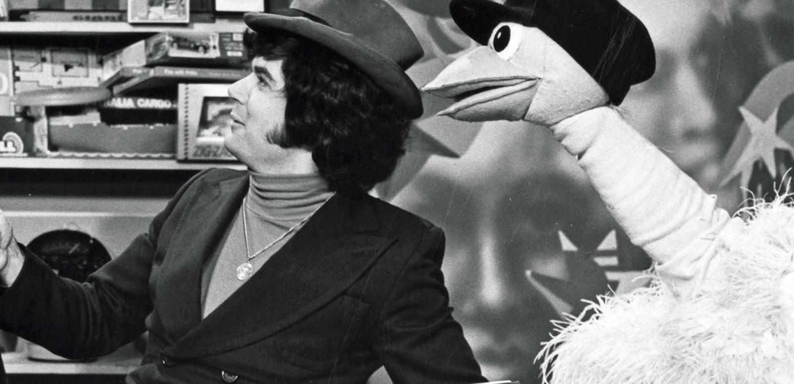 A man wearing a hat sitting next to an ostrich puppet who is also wearing a hat. They are both turned to their right.